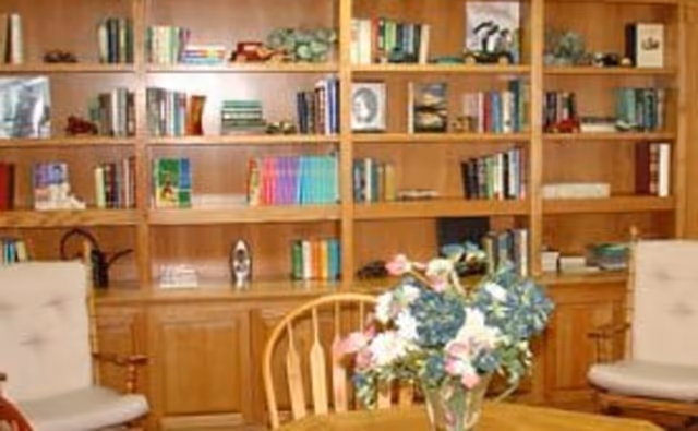 A cozy room with wooden bookshelves filled with books and decorative items. In front of the shelves is a round wooden table with a vase of colorful flowers. Two cushioned armchairs are positioned on either side of the table.