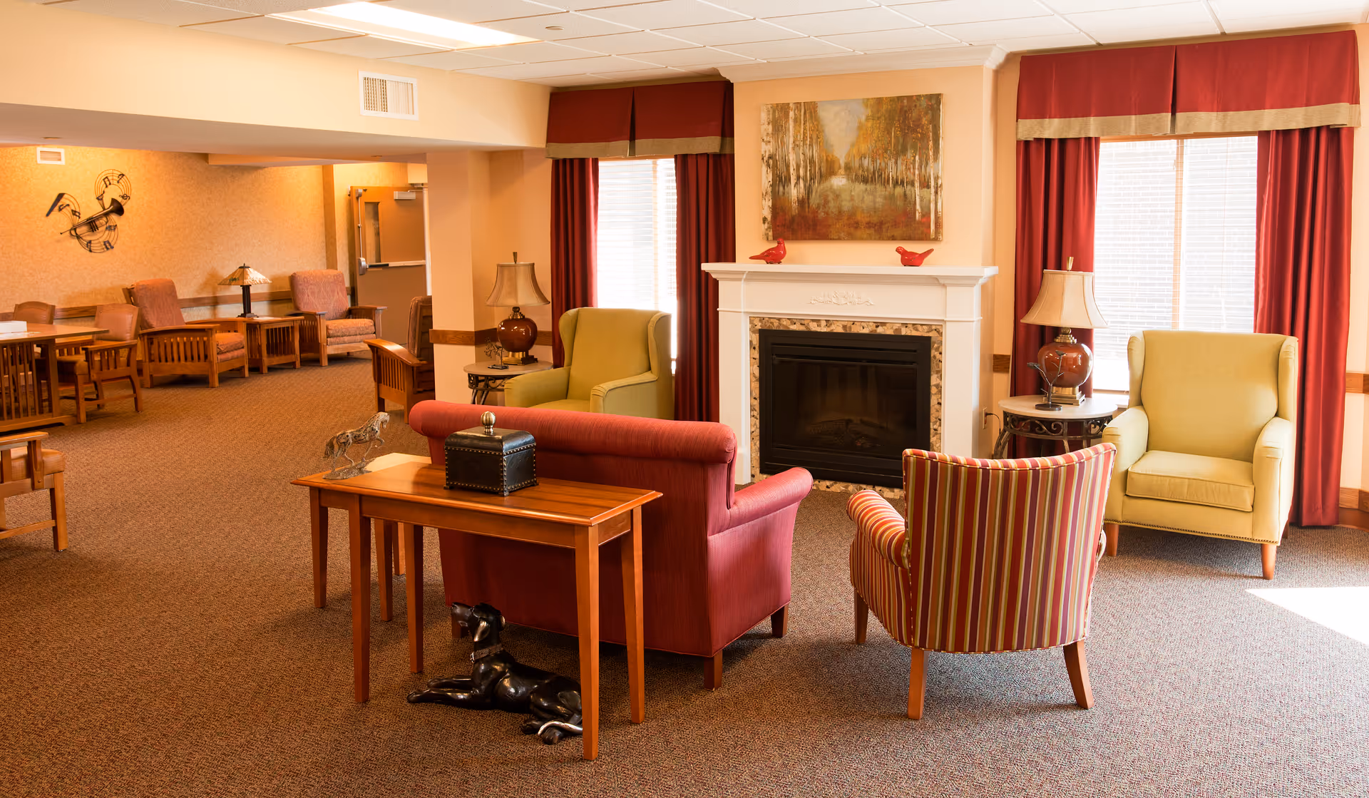 A cozy senior living center common area with a fireplace, surrounded by various upholstered chairs including a red loveseat, a striped armchair, and two green armchairs. There are side tables with lamps, red curtains on the windows, and a painting above the fireplace. Additional seating and tables are visible in the background.