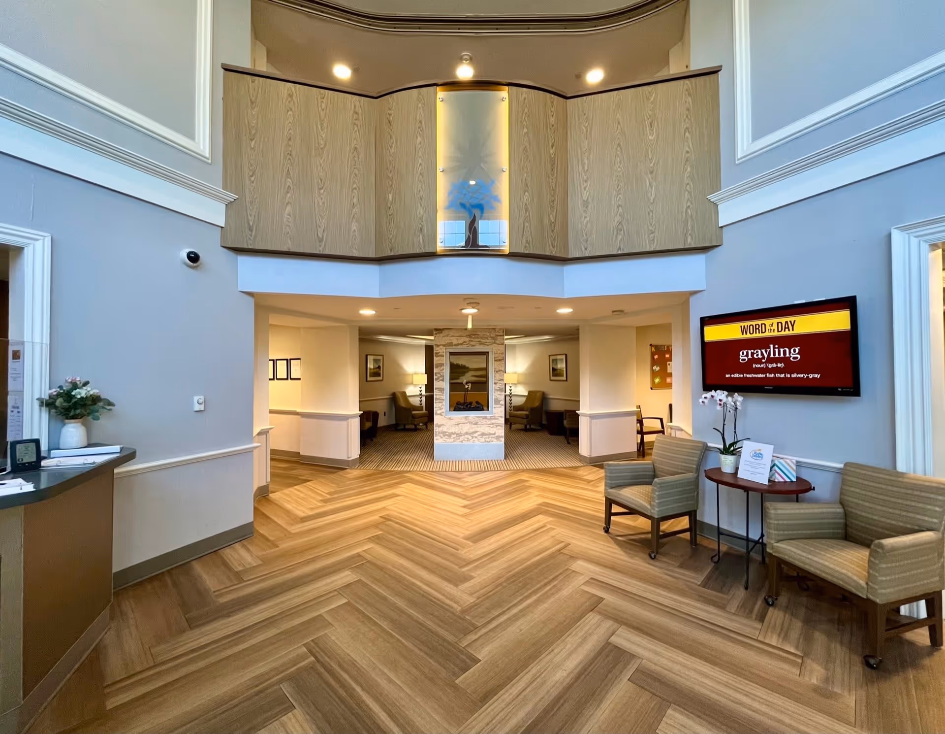 Interior view of a senior living facility lobby area with wood-patterned herringbone flooring, a central fireplace, seating areas with chairs, a small table with a plant and informational materials, and a wall-mounted screen displaying a word of the day. The space has high ceilings and a balcony with wood paneling above.