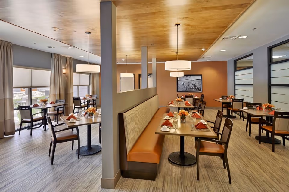 A bright and modern dining room with multiple tables set for meals. The tables have orange napkins, glassware, and small floral centerpieces. There is a long cushioned bench along a partial wall dividing the room, with wooden chairs on the opposite side of the tables. Large windows with curtains allow natural light to fill the space, and pendant lights hang from a wooden ceiling.