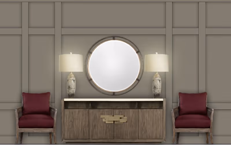 A symmetrical interior setup featuring a wooden sideboard with a white countertop, a round mirror mounted above it, and two matching table lamps with beige shades on either side. Flanking the sideboard are two wooden armchairs with dark red cushions. The background wall has a paneled design painted in a neutral gray tone.