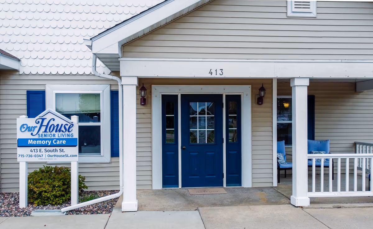 Front entrance of Our House Senior Living - Rice Lake Memory Care facility with a blue door, white pillars, and a sign displaying the facility name, address, phone number, and website.
