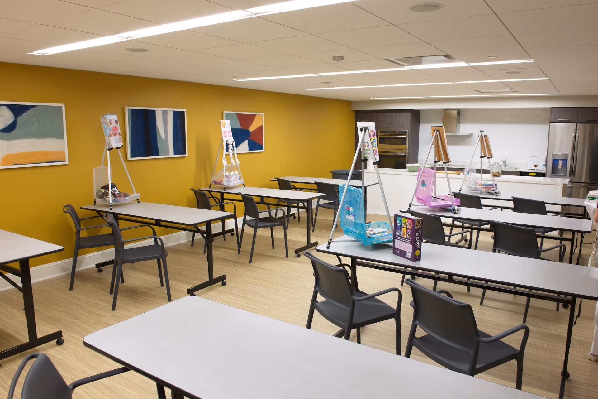 A bright room with multiple tables and chairs arranged in rows, each table equipped with an easel holding art supplies. The walls are painted mustard yellow and decorated with abstract paintings. In the background, there is a kitchen area with modern appliances including an oven and refrigerator.