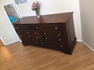A dark wooden dresser with multiple drawers, placed against a beige wall on a light wooden floor. On top of the dresser is a vase with pink flowers. A mirror is partially visible on the left side of the image.