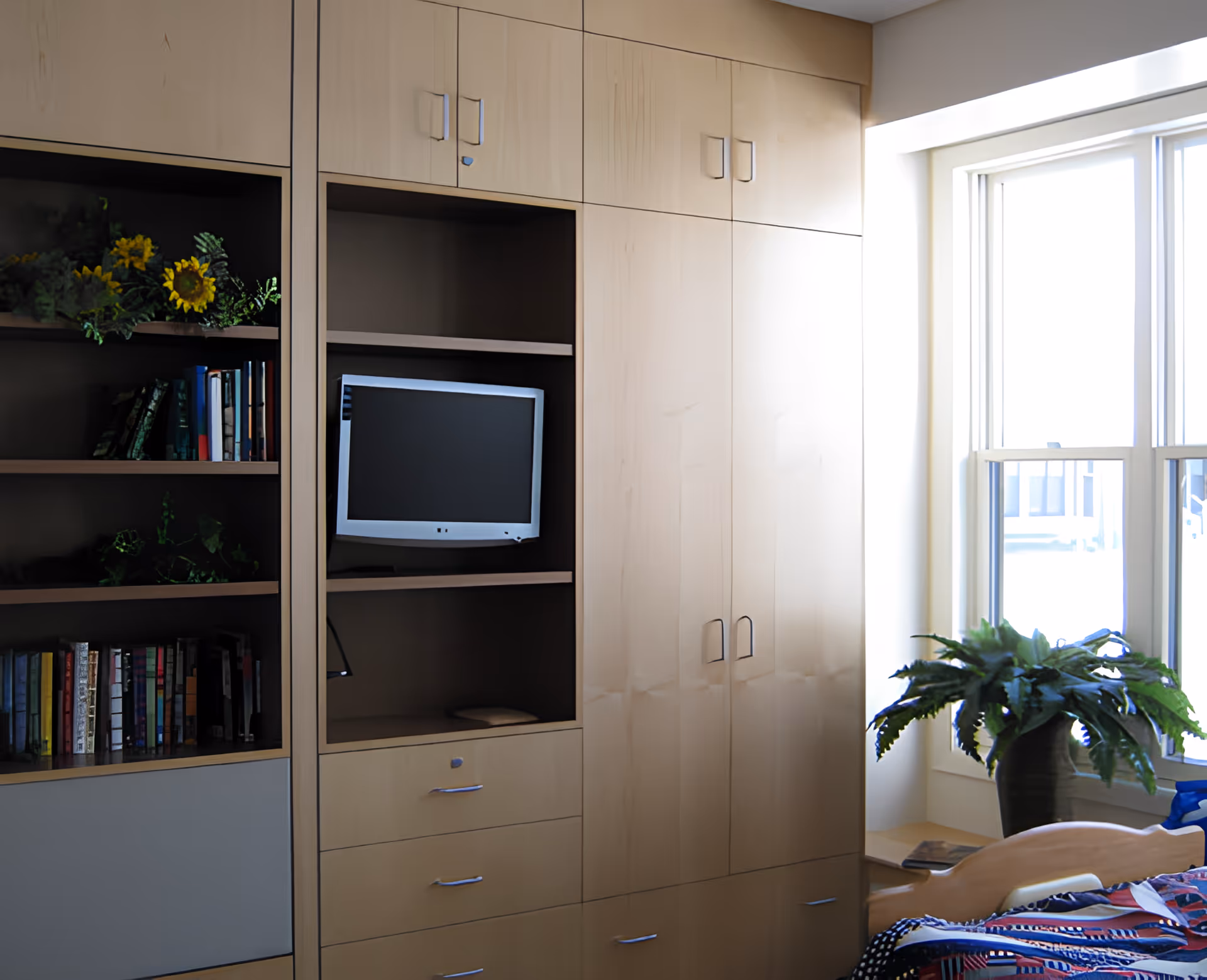 A cozy bedroom corner with built-in wooden cabinets and shelves. The shelves hold books and decorative plants, and there is a small flat-screen TV mounted in one of the open cabinet spaces. A window with white trim lets in natural light, and a potted plant sits on the windowsill next to a bed with a colorful quilt.