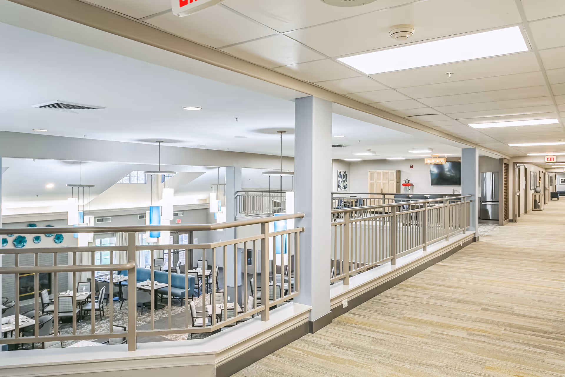 Interior hallway overlooking a bright communal dining and lounge area with railings, tables, and seating.