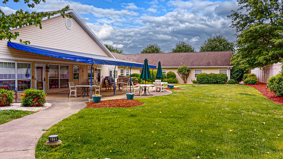 Outdoor patio area of a senior living facility with tables, chairs, and umbrellas on a concrete patio next to a beige building with a blue awning. The area is surrounded by green grass, shrubs, and trees under a partly cloudy sky.
