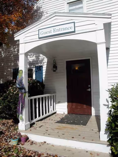 White building entrance with a covered porch, 'Guest Entrance' sign, and a life-size figure near railing.
