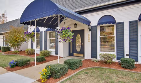 Front entrance of a light-colored building with a blue fabric awning, hanging flower baskets, and landscaped shrubs along a walkway.
