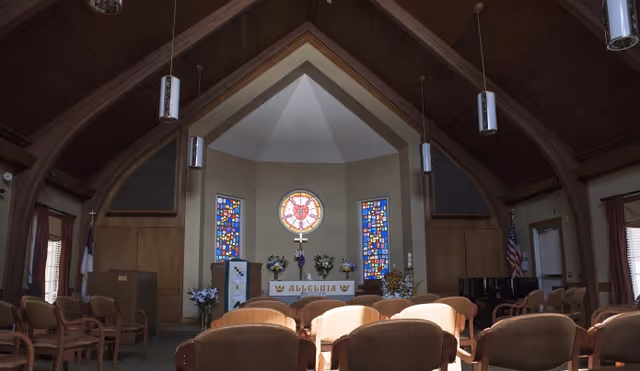 Interior view of a chapel with wooden vaulted ceiling, stained glass windows, an altar with flowers and a cross, and rows of chairs facing the altar.