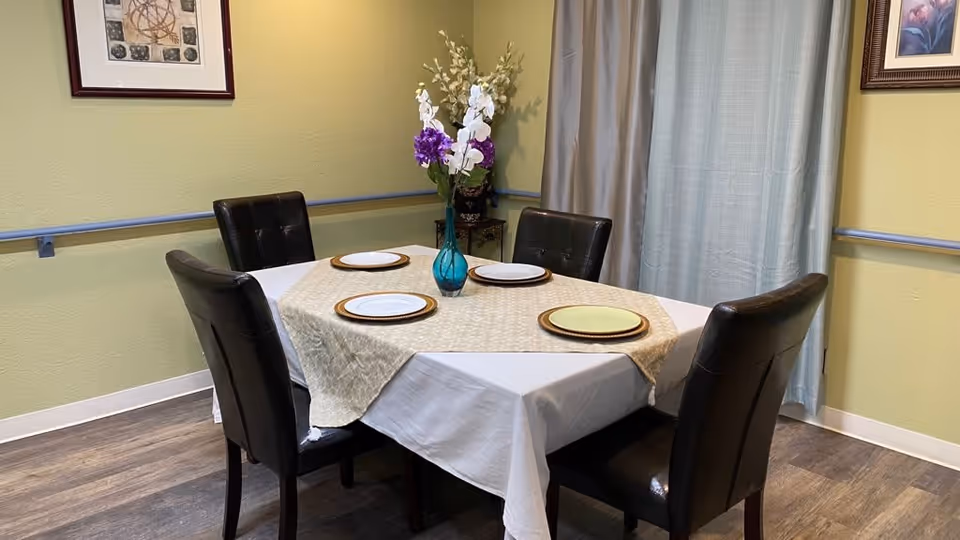 A dining area with a rectangular table covered with a white tablecloth and a beige table runner. Four dark brown leather chairs surround the table. On the table, there are four plates and a blue vase with white and purple flowers. The room has light green walls, a framed picture on the wall, a curtain covering a window, and wood-style flooring.