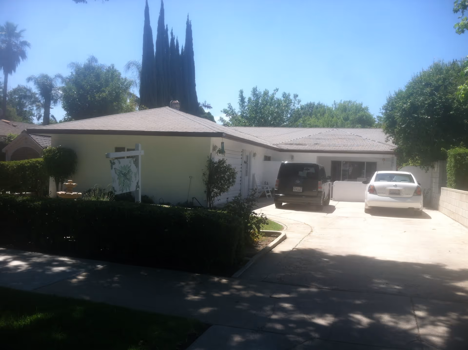 Exterior view of a single-story residential building with a driveway containing two parked cars, surrounded by trees and bushes under a clear blue sky.