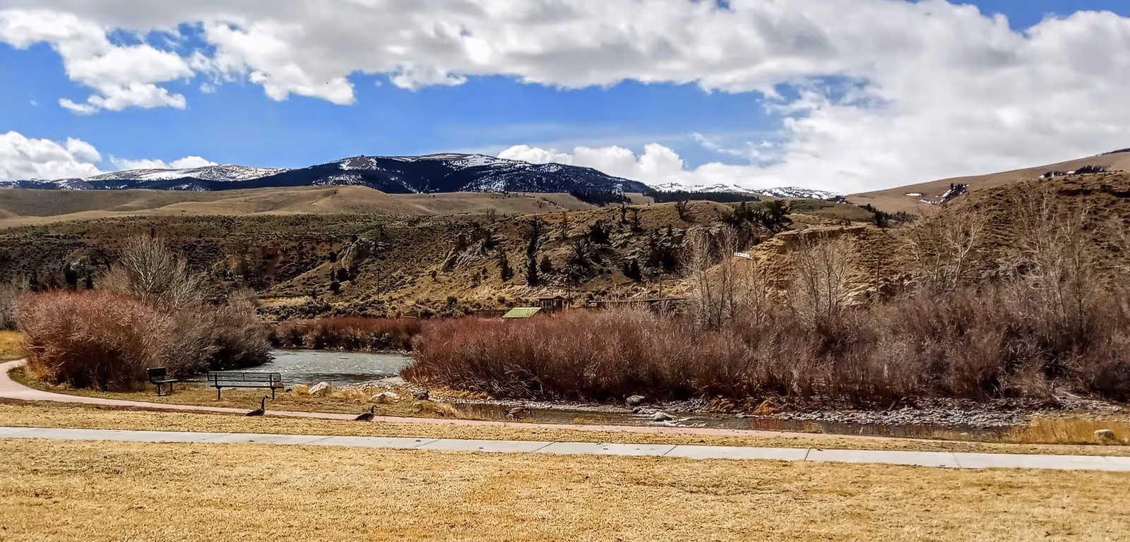 A grassy park area with a walking path and bench beside a small river, shrubbery and hills, and snow-capped mountains under a partly cloudy sky.