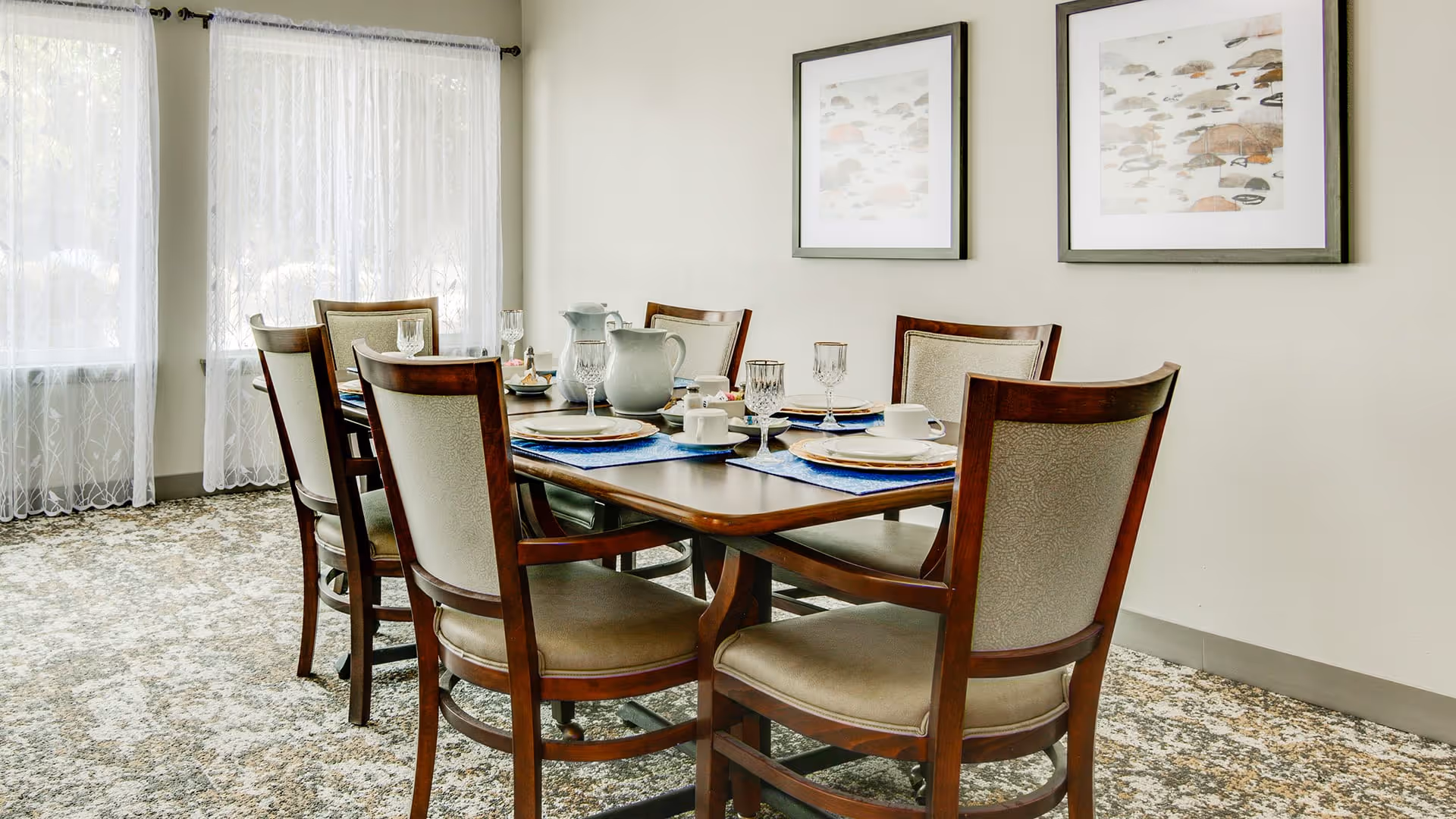 A dining room with a wooden table set for six people. The table has plates, glasses, cups, and a pitcher. There are six wooden chairs with cushioned seats around the table. The room has light-colored walls with two framed pictures hanging on one wall and large windows with sheer white curtains allowing natural light to enter.