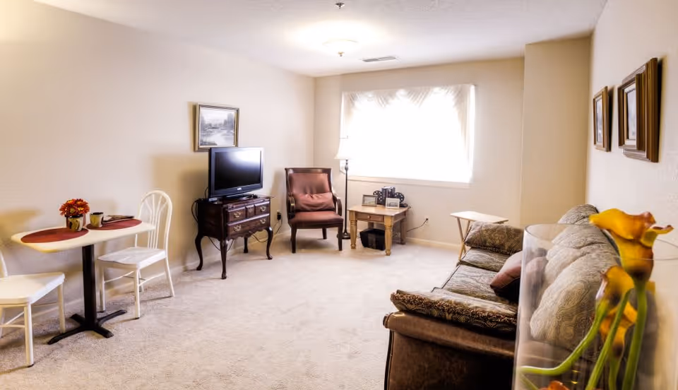 A cozy living room with a beige carpet and cream-colored walls. The room features a small dining table with two white chairs and a red placemat with a flower vase. There is a wooden TV stand with a flat-screen TV, a cushioned armchair, and a side table with framed photos and a lamp near a window with sheer curtains. A patterned sofa with cushions is on the right side, and a vase with yellow flowers is in the foreground.