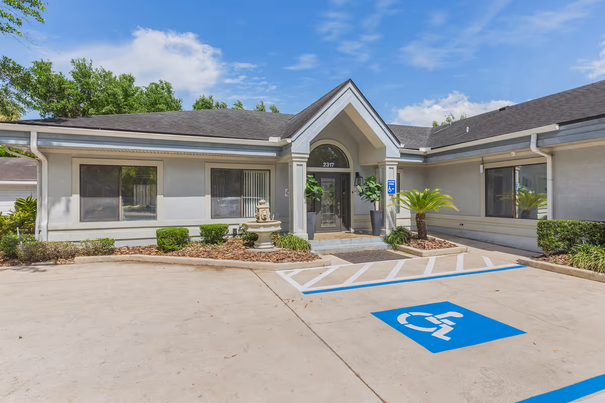 Exterior front entrance of a single-story senior living facility building with a covered porch, large windows, and a handicapped parking space in front. The building is surrounded by small shrubs and plants under a partly cloudy blue sky.