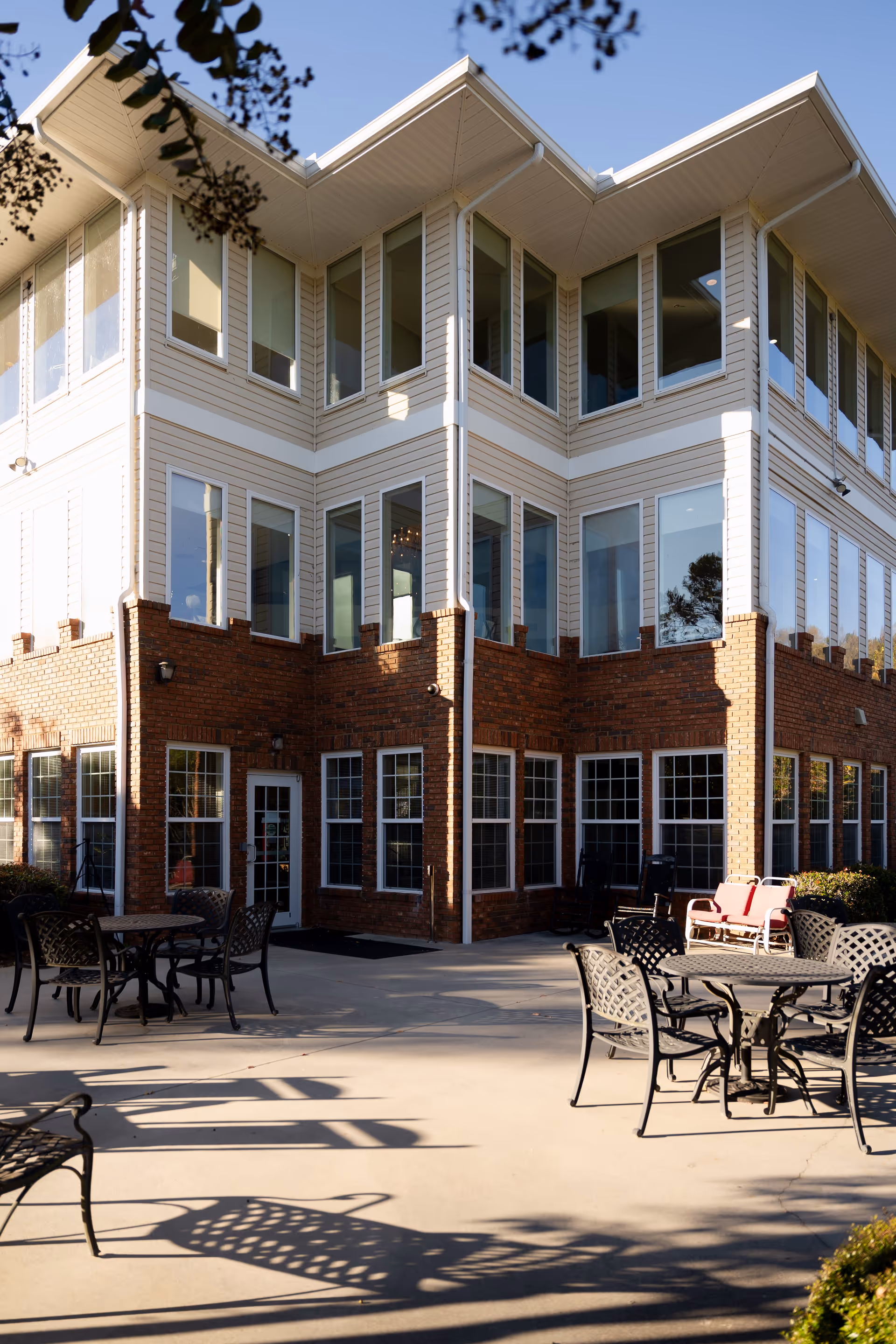 Outdoor patio area of a senior living facility with metal tables and chairs arranged on a concrete surface. The building has a brick lower level and beige siding on the upper level with many large windows. Some greenery and tree branches are visible around the patio.