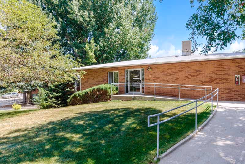 Single-story brick building front with a wheelchair ramp, glass entrance doors, and a grassy lawn with trees.