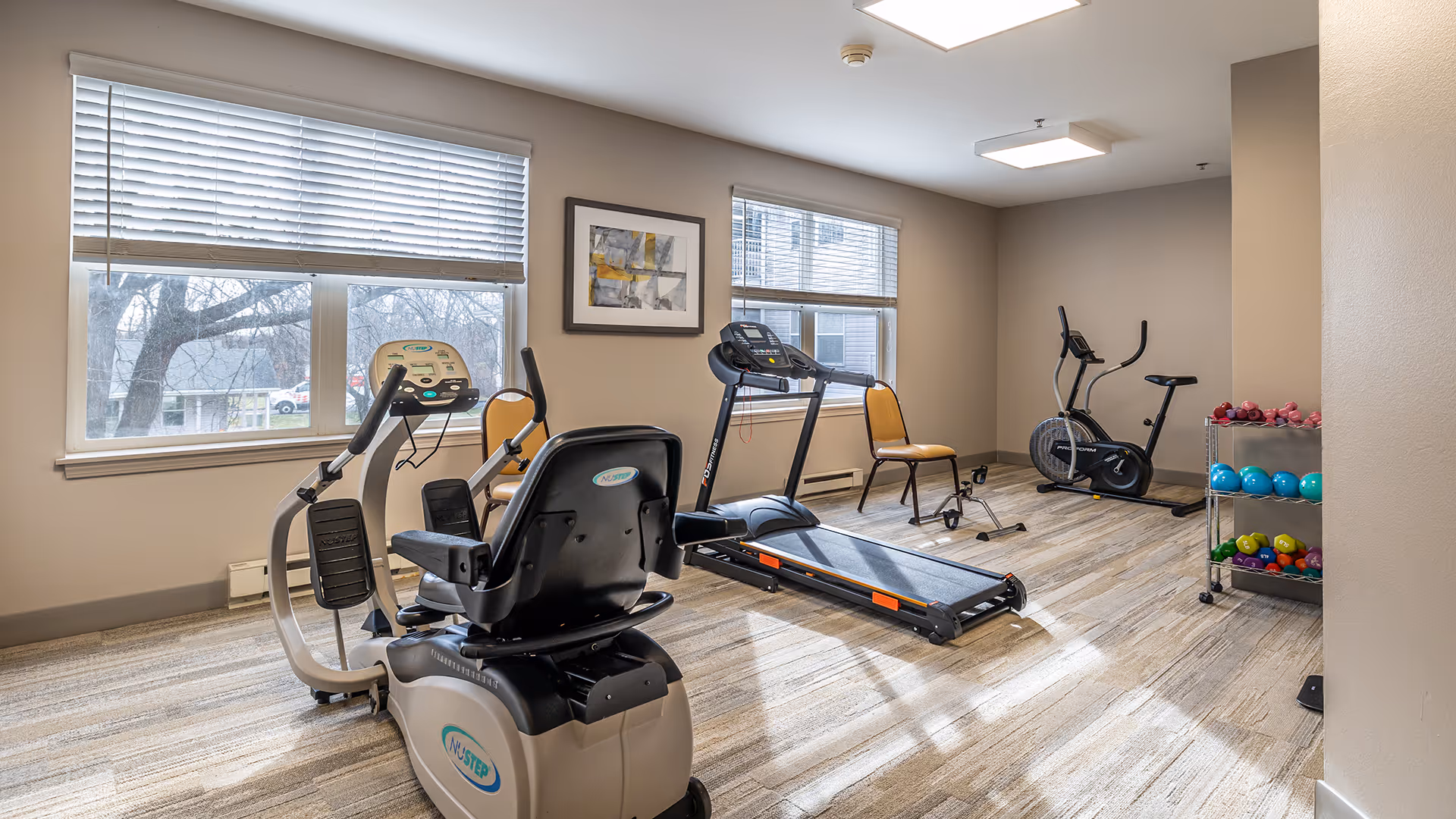 A small fitness room with exercise equipment including a recumbent bike, treadmill, stationary bike, and a rack of colorful dumbbells. The room has large windows with blinds, light-colored walls, and wood-patterned flooring. Two yellow chairs are placed near the windows.