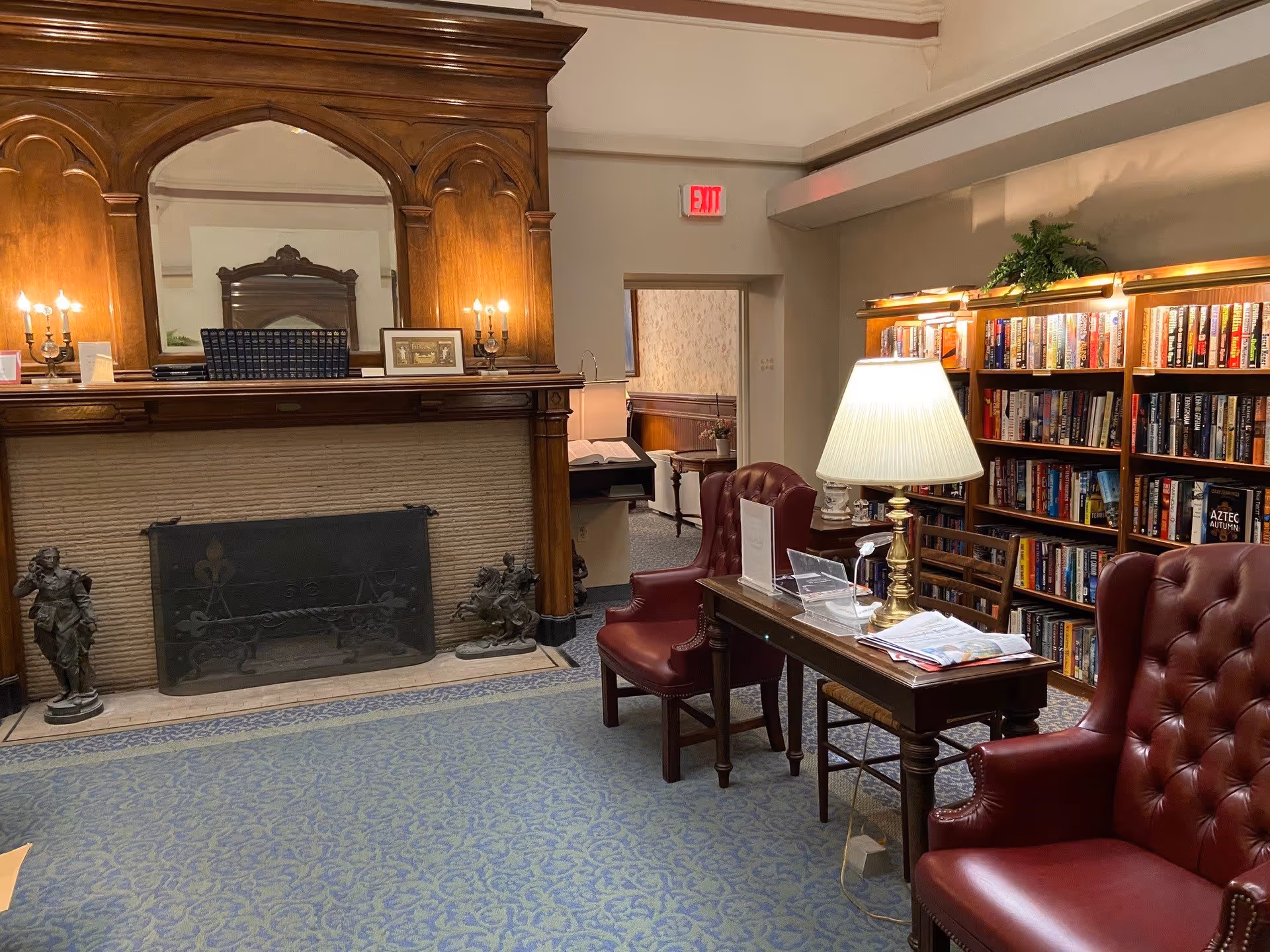 A cozy library room with a large wooden fireplace mantel, two red leather armchairs, a wooden table with a lamp and reading materials, and bookshelves filled with books along the wall. There is an exit sign above a doorway in the background.