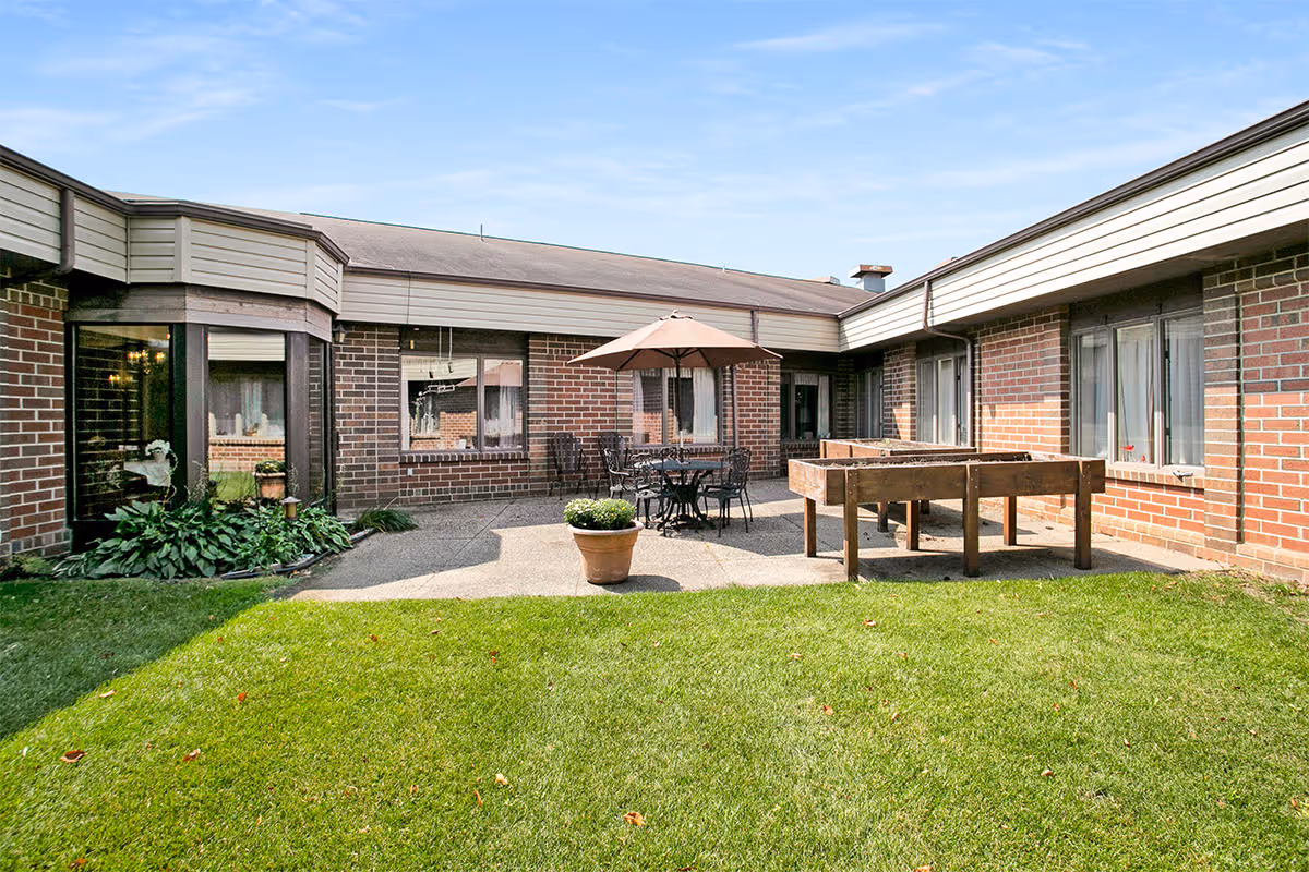 Outdoor courtyard area of a senior living facility with a green lawn, a concrete patio, a round table with chairs and an umbrella, two raised garden beds, and brick building walls with windows surrounding the space.