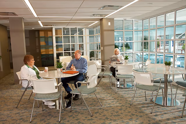 A bright common area in a senior living facility with several round tables and chairs. Three elderly people are seated at two tables; one woman is holding a cup, a man is sitting with a folder on the table, and another woman is reading a newspaper. Large windows reveal an indoor swimming pool area outside the room.