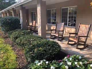 Outdoor patio area of a senior living facility with several wooden rocking chairs lined up along the covered porch. There are bushes and flowering plants in the foreground, and the building has beige siding with multiple windows.