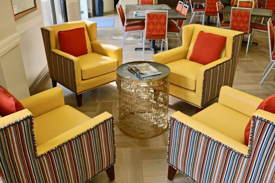 Four yellow armchairs with striped sides and red cushions arranged around a round decorative metal table in a communal seating area with dining chairs in the background.