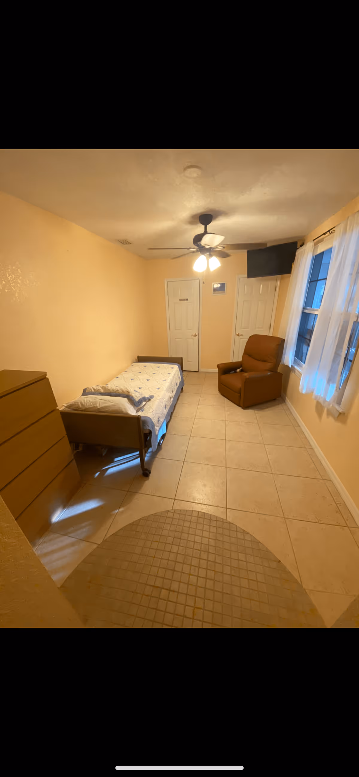 A small, simple bedroom with beige walls and tiled floor. The room contains a single bed with a white mattress and pillow, a brown recliner chair near two windows with white curtains, a wooden dresser, and a ceiling fan with lights. There are two closed white doors on the far wall.