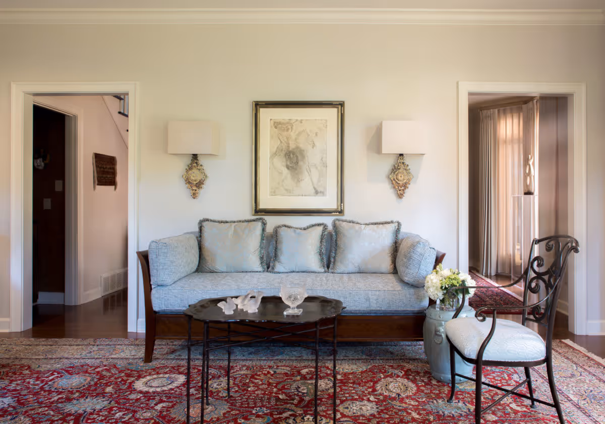A cozy living room with a light blue cushioned sofa adorned with matching pillows, a decorative framed artwork above it, and two wall-mounted lamps on either side. In front of the sofa is a small black coffee table with glass decor items. To the right, there is a black metal chair with a white cushion and a large vase with white flowers. The floor is covered with a red patterned rug, and there are two doorways visible on either side of the sofa leading to other rooms.