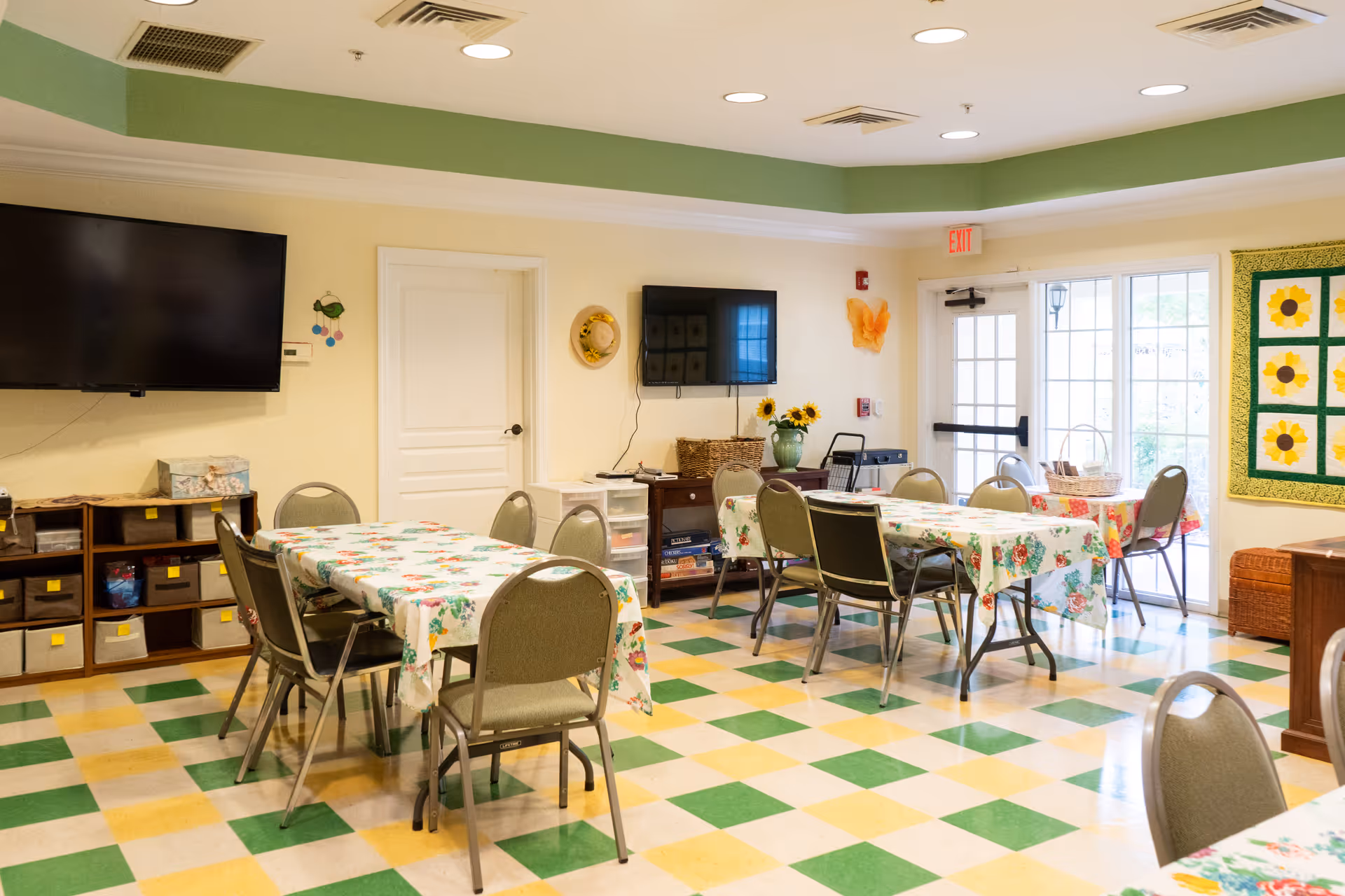 A bright and cheerful common area with tables covered in floral tablecloths and surrounded by chairs. The room has a checkered green, yellow, and white floor, two wall-mounted flat-screen TVs, a door, large windows letting in natural light, and sunflower-themed decorations including a quilt on the wall and a vase with sunflowers on a table.