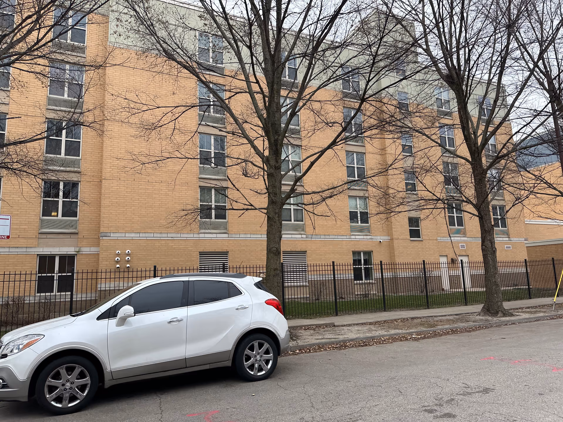 Street-facing view of a multi-story tan brick residential building with leafless trees and a white SUV parked in front.