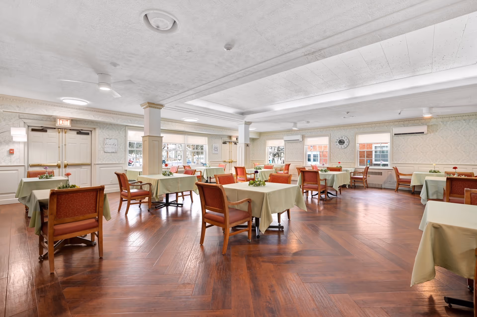 A spacious dining room with multiple tables covered in light green tablecloths, each table surrounded by wooden chairs with red cushions. The room has large windows letting in natural light, a wooden floor with a herringbone pattern, and light-colored walls with decorative molding. There are small flower arrangements on each table and ceiling lights providing additional illumination.