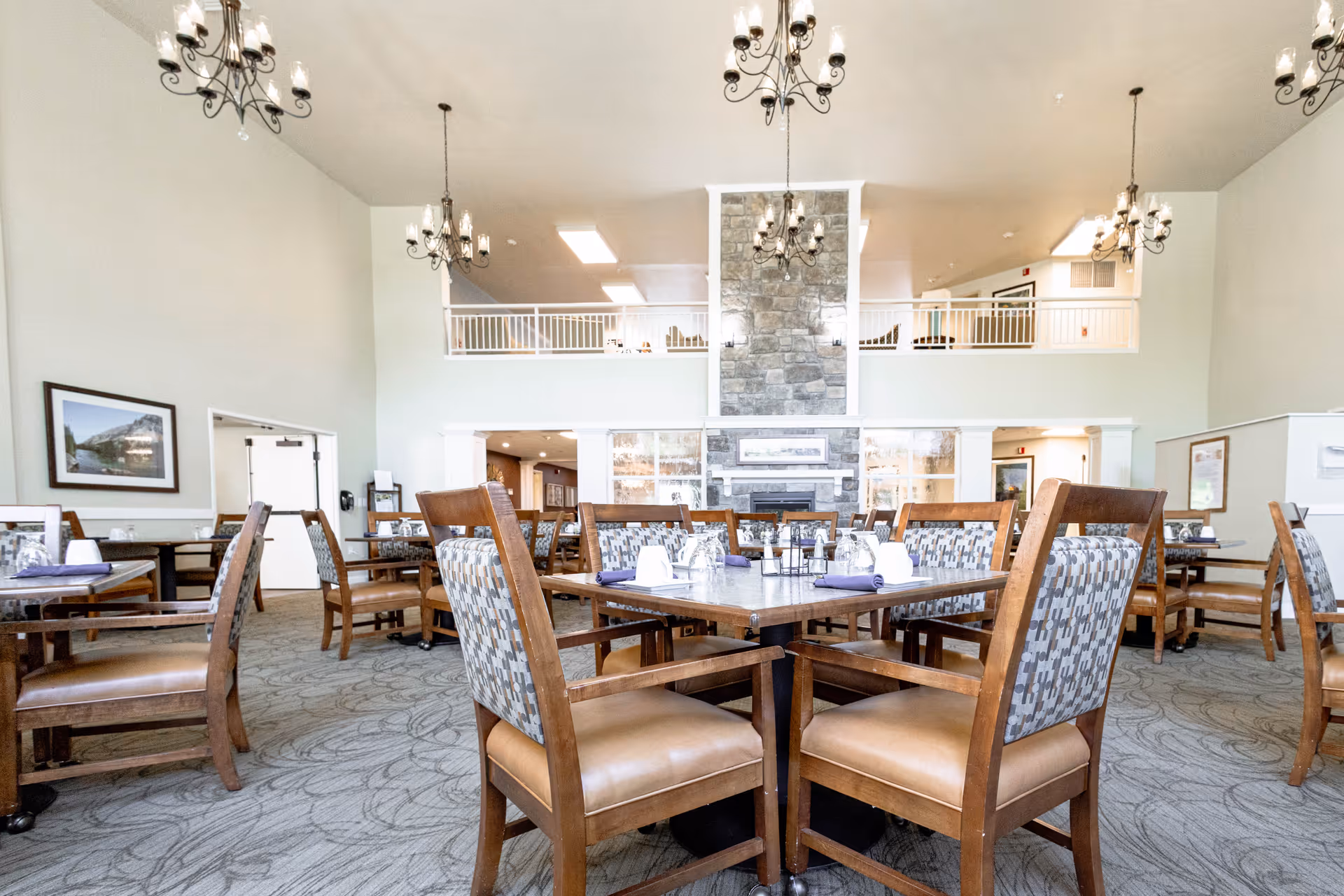A spacious dining room with multiple wooden tables and chairs arranged neatly. Each table is set with glasses, napkins, and condiments. The room features a high ceiling with elegant chandeliers and a large stone fireplace in the center background. There is a balcony railing above the fireplace area and framed artwork on the walls.