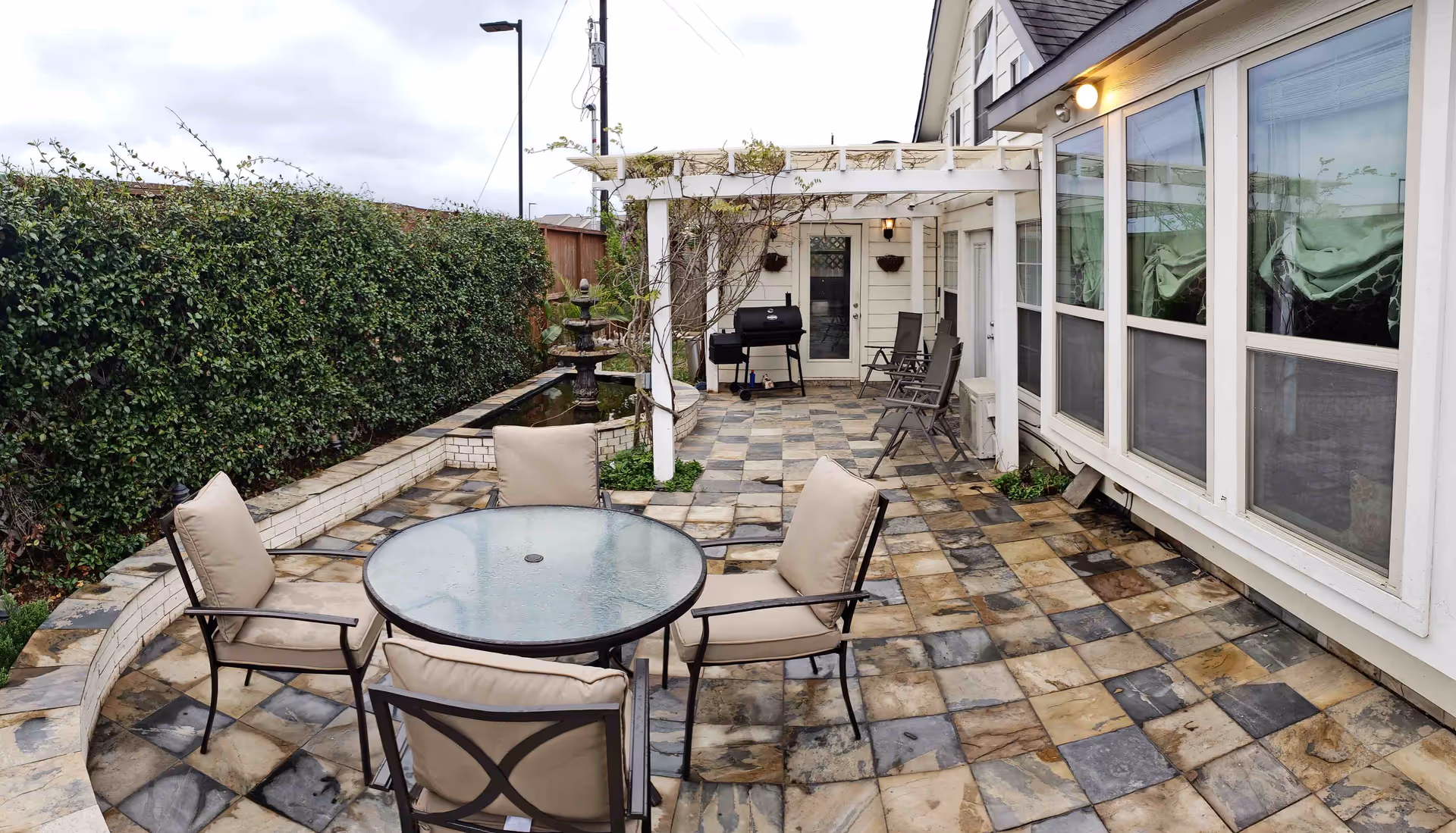 Outdoor patio area with a round glass table and four cushioned chairs on a tiled floor. There is a white pergola with some climbing plants, a black grill, and additional chairs along the side of a white building with large windows. A hedge runs along the left side of the patio.