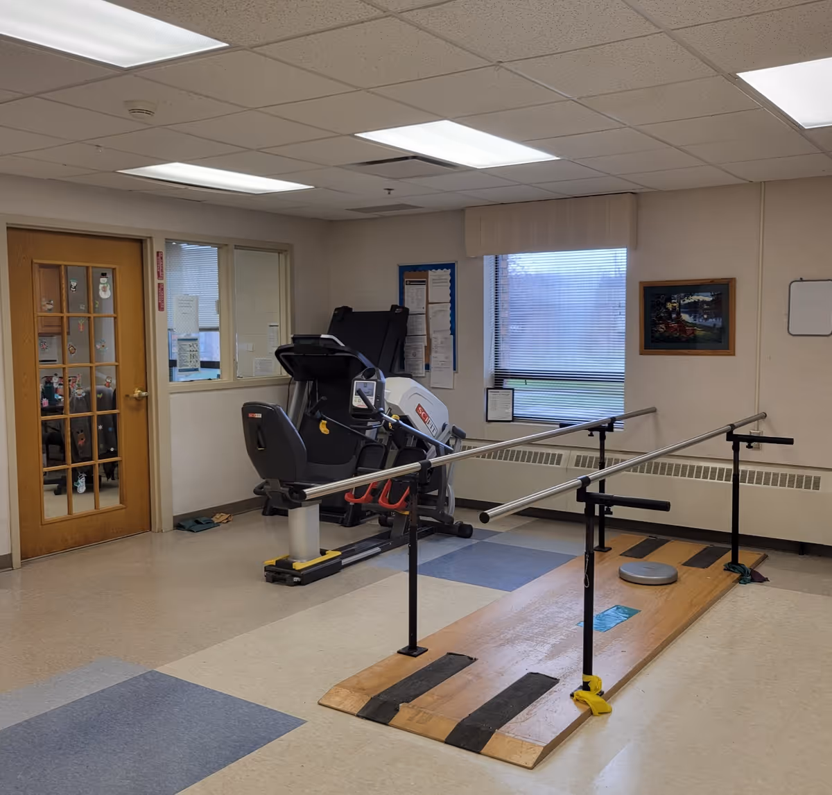 A rehabilitation room with parallel bars for walking exercises on a wooden platform, a recumbent exercise bike, a treadmill, a window with blinds, a bulletin board, a framed picture on the wall, and a wooden door with glass panels.