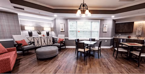 A spacious common area with wooden flooring featuring two seating areas with chairs and a red sofa, a round ottoman, two small dining tables with chairs, decorative lamps, and wall art. Large windows with blinds allow natural light into the room.