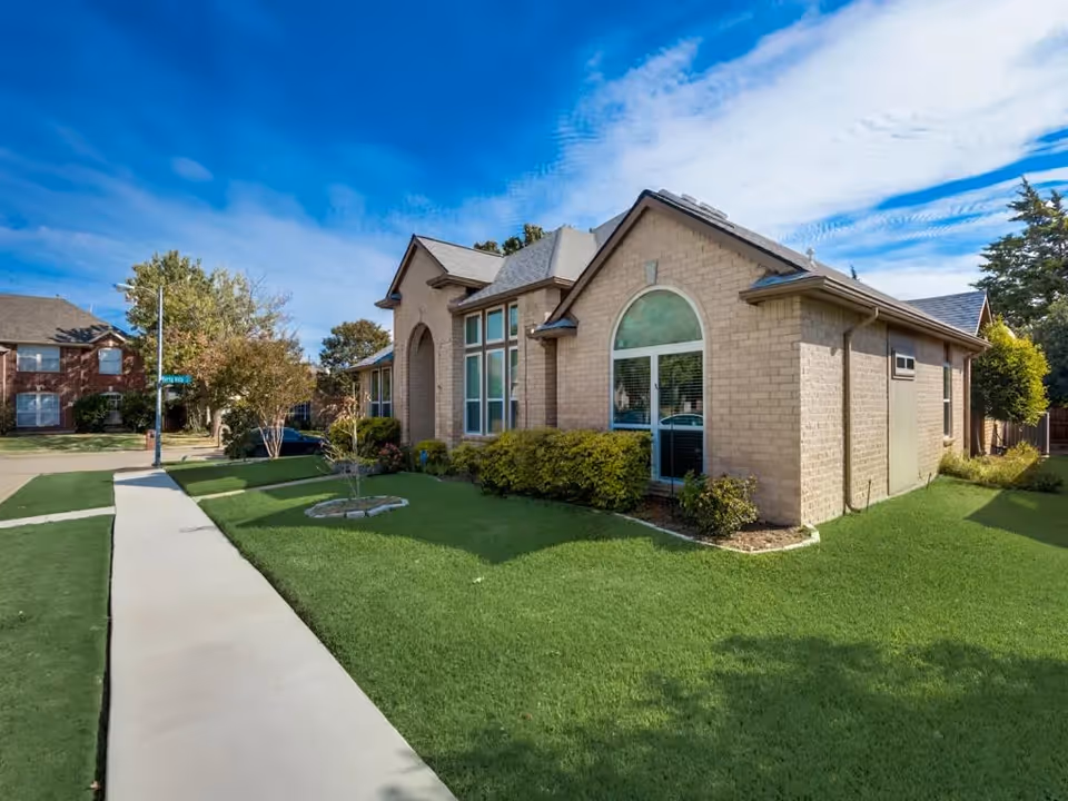 Exterior view of a single-story brick building with large windows and a well-maintained green lawn under a blue sky with scattered clouds. A sidewalk runs alongside the building, and there are trees and shrubs around the property.