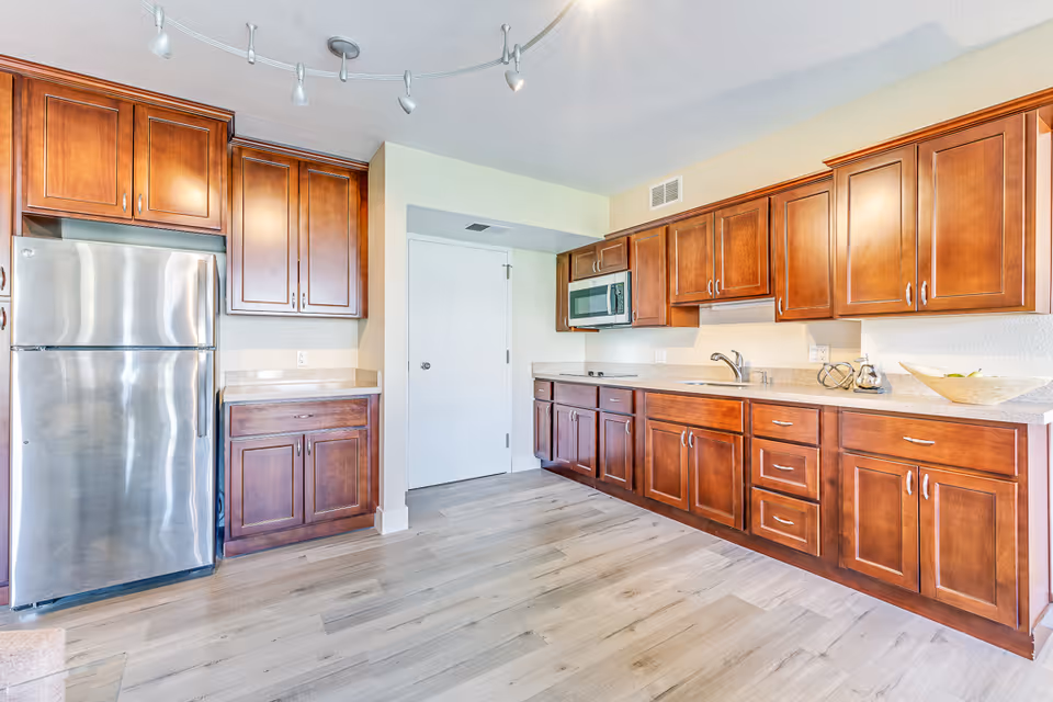 A bright kitchen with wooden cabinets, a stainless steel refrigerator, a built-in microwave, a sink, and light-colored countertops. The floor is light wood, and there is a white door in the background.