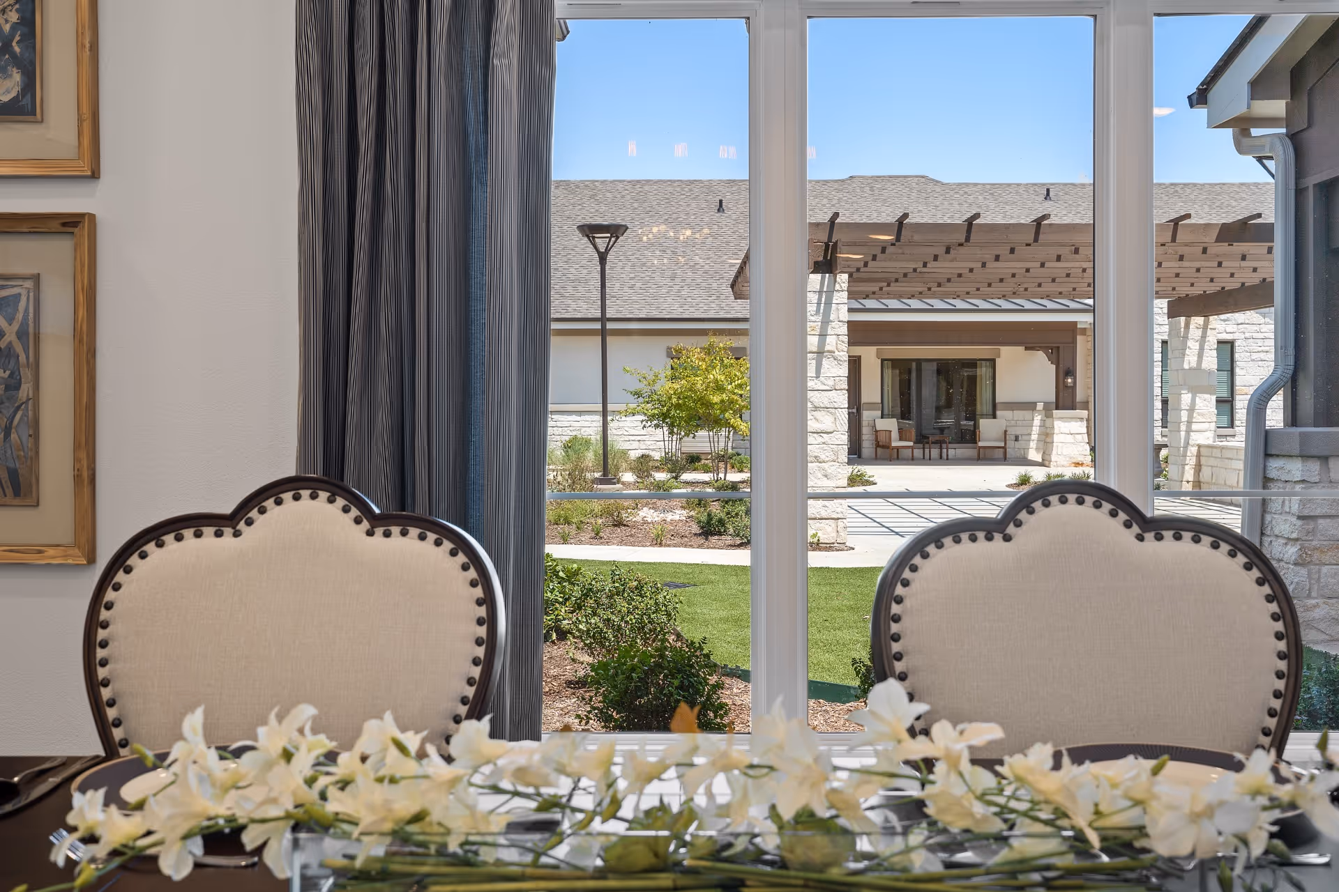 View from inside a dining room showing two upholstered chairs with nailhead trim in front of a large window. Outside the window, there is a landscaped garden area with bushes, a lamp post, and a building with stone pillars and a pergola under a clear blue sky. White flowers are arranged on the dining table in the foreground.