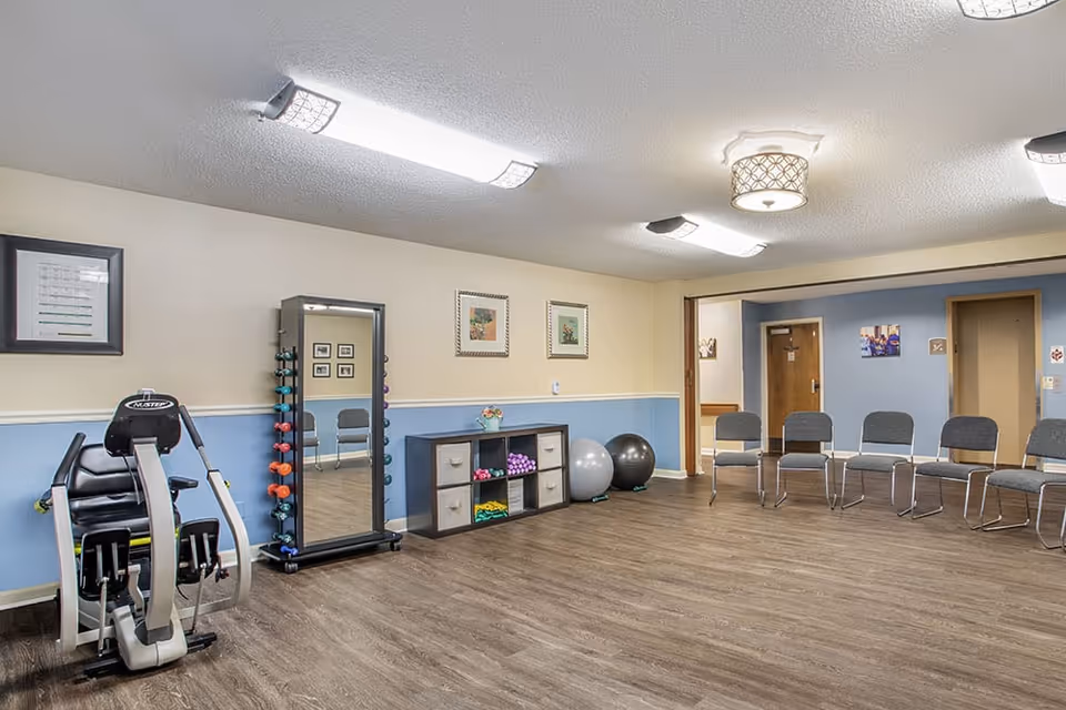An indoor activity/exercise room with chairs along the wall, a piece of fitness equipment, free weights on a mirror rack, and exercise balls.