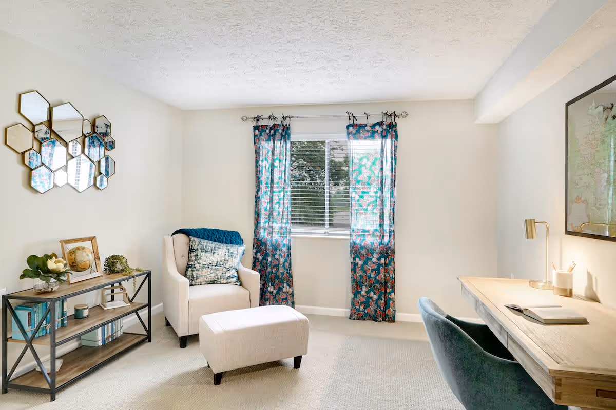 A bright and cozy room with a beige armchair and matching ottoman placed near a window with blue patterned curtains. To the left, there is a metal and wood shelf holding books, a globe, and decorative items. On the right side, there is a wooden desk with a blue chair, a lamp, and an open book. The walls are light-colored, and a decorative hexagonal mirror is hung on the left wall.