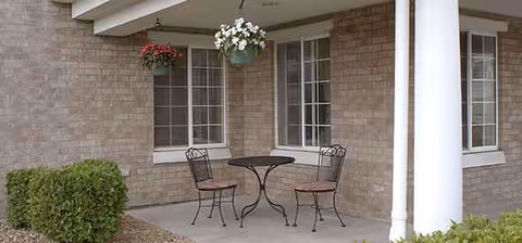 Outdoor covered patio with a round table, two chairs, and hanging baskets against brick walls.