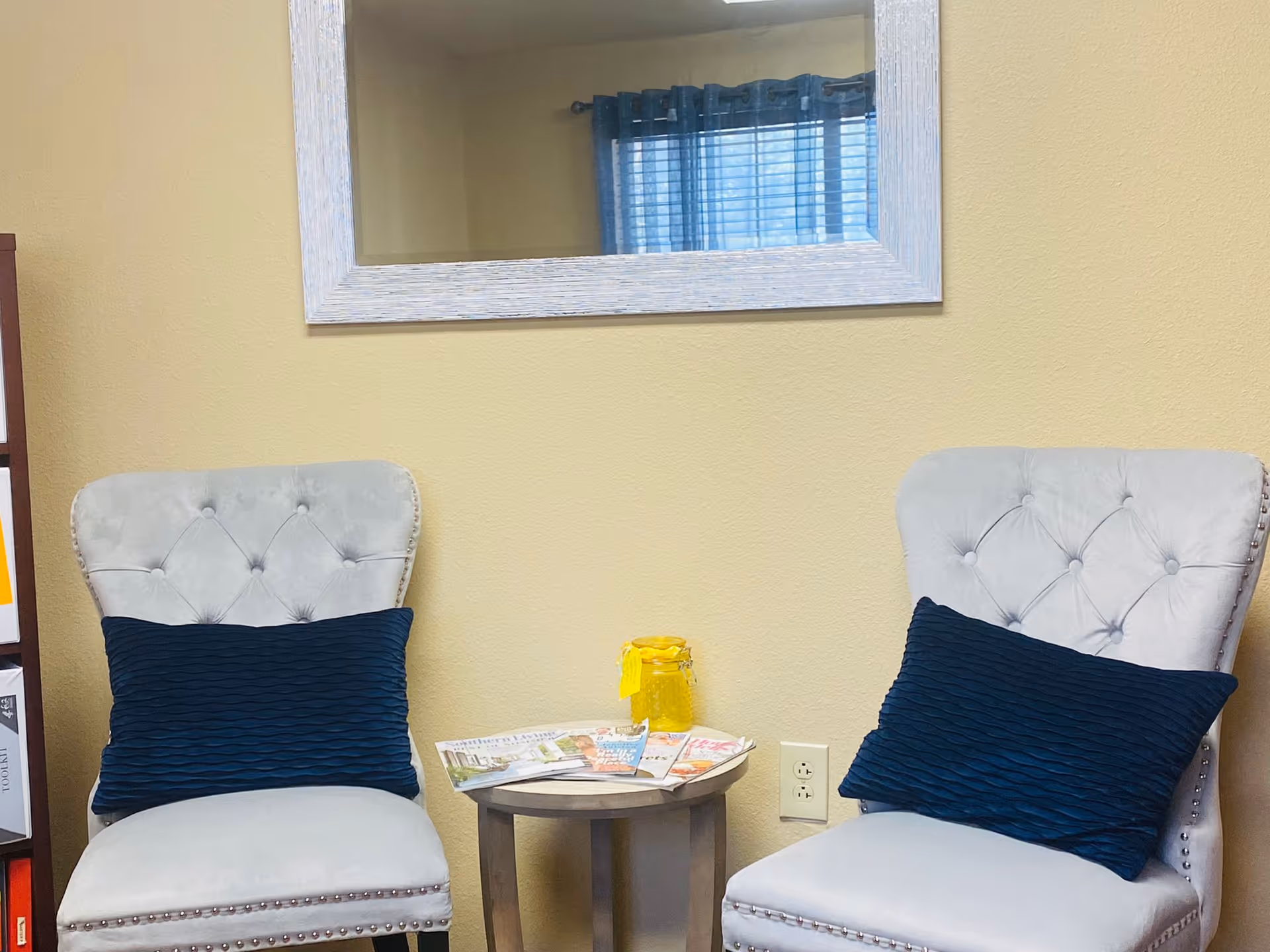Two tufted upholstered chairs with blue pillows flank a small round table holding magazines and a jar beneath a wall mirror in a cozy seating area.