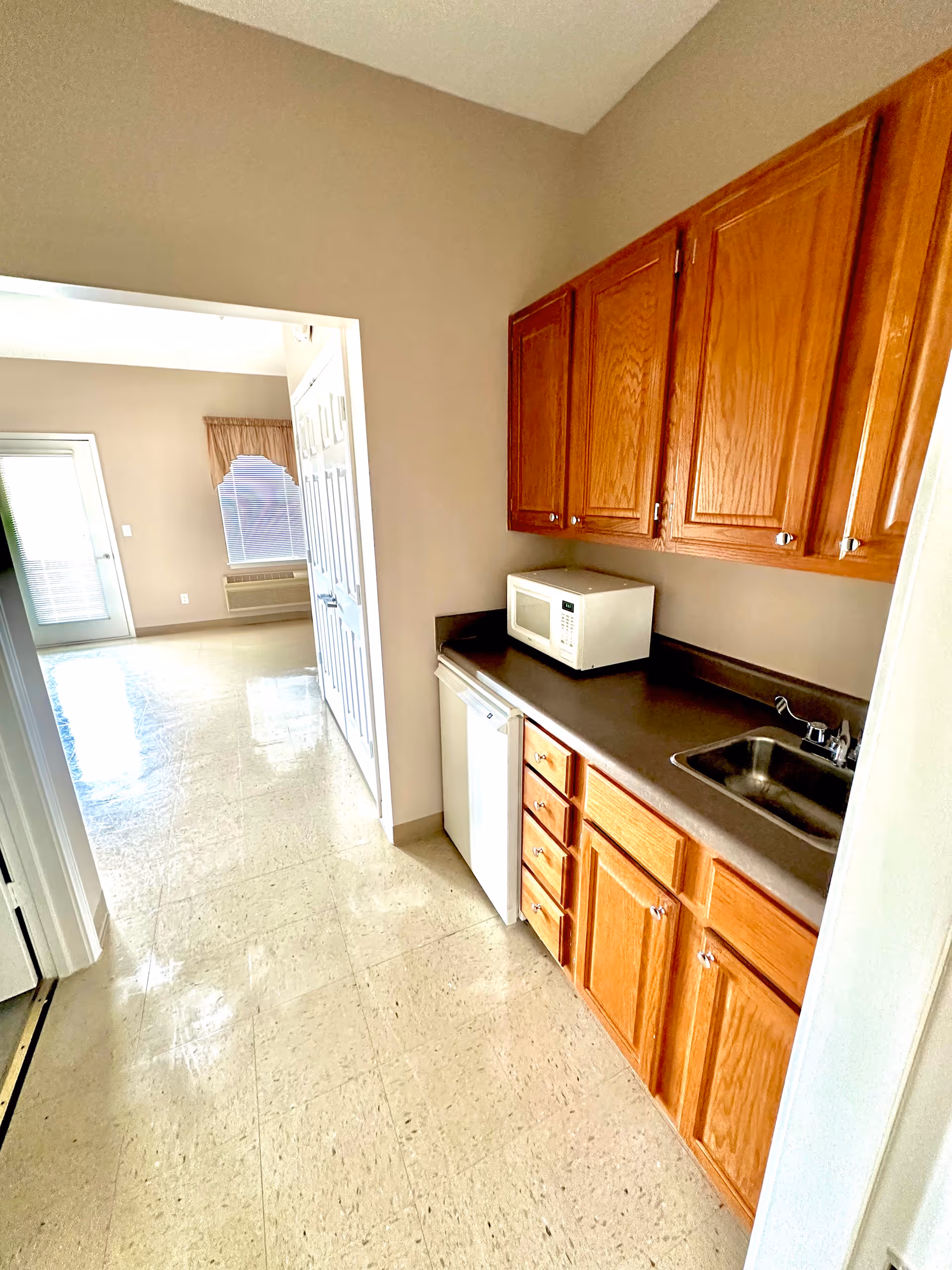 Small kitchenette with wooden cabinets, countertop, microwave, mini-fridge and sink opening into a bright living area.