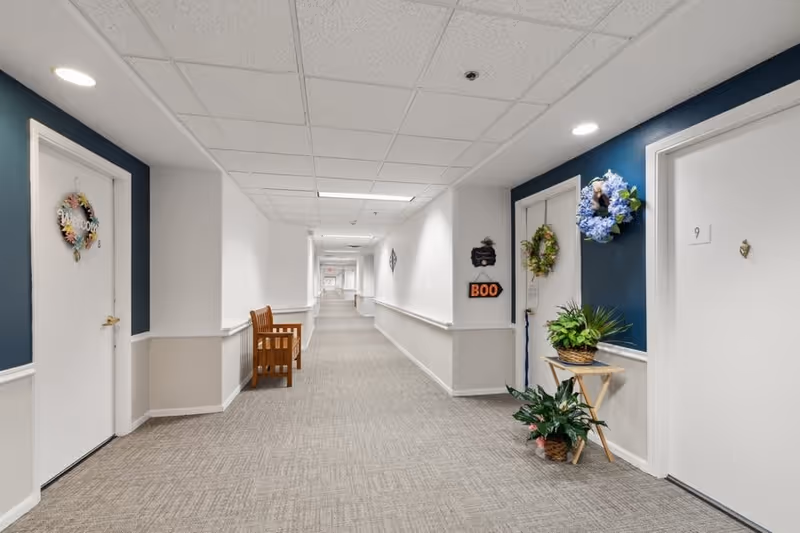 A long, well-lit hallway in a senior living facility with white walls and blue accent panels. Several doors line the hallway, each decorated with wreaths and signs. A wooden bench is placed along one wall, and there are potted plants on a small table near one of the doors. The carpet is light gray with a subtle pattern, and the ceiling has recessed lighting.