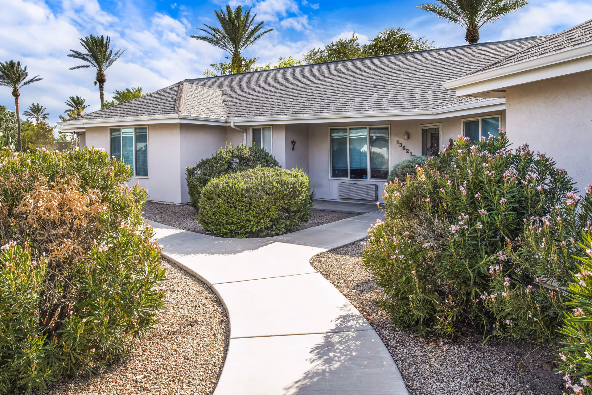Exterior view of a single-story senior living community building with a gray shingled roof, beige walls, and several windows. A curved concrete walkway leads to the entrance, surrounded by well-maintained bushes and desert landscaping with gravel. Palm trees and a partly cloudy blue sky are visible in the background.