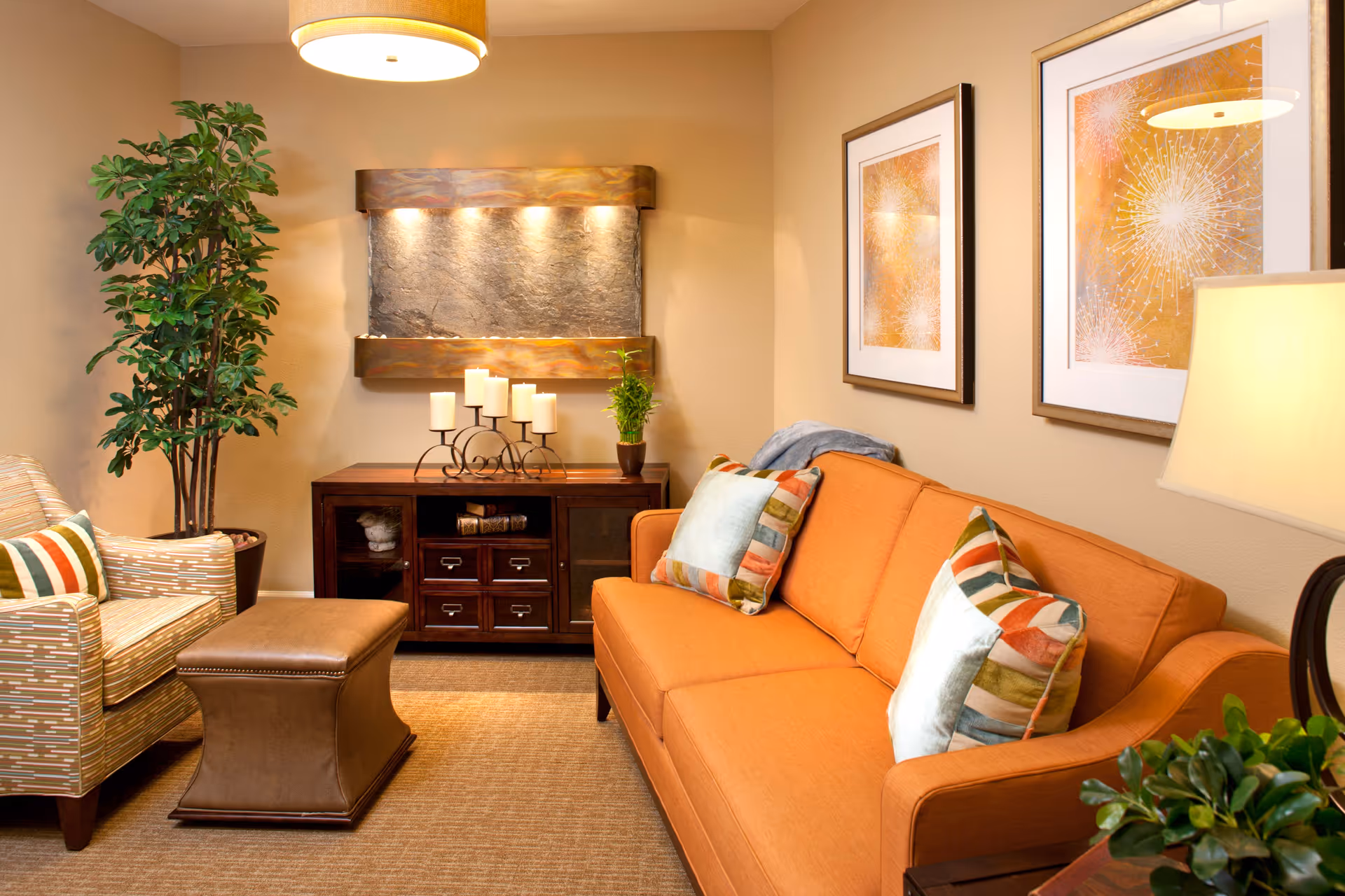 A cozy living room with an orange sofa adorned with colorful striped pillows, a patterned armchair, a brown leather ottoman, a wooden cabinet with candles and a small plant on top, two framed abstract artworks on the wall, a tall green plant in the corner, and a warm overhead light fixture.