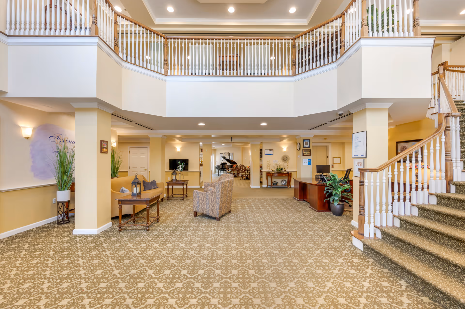 Spacious two-story senior living lobby with seating areas, a reception desk, carpeted staircase and balcony railing.