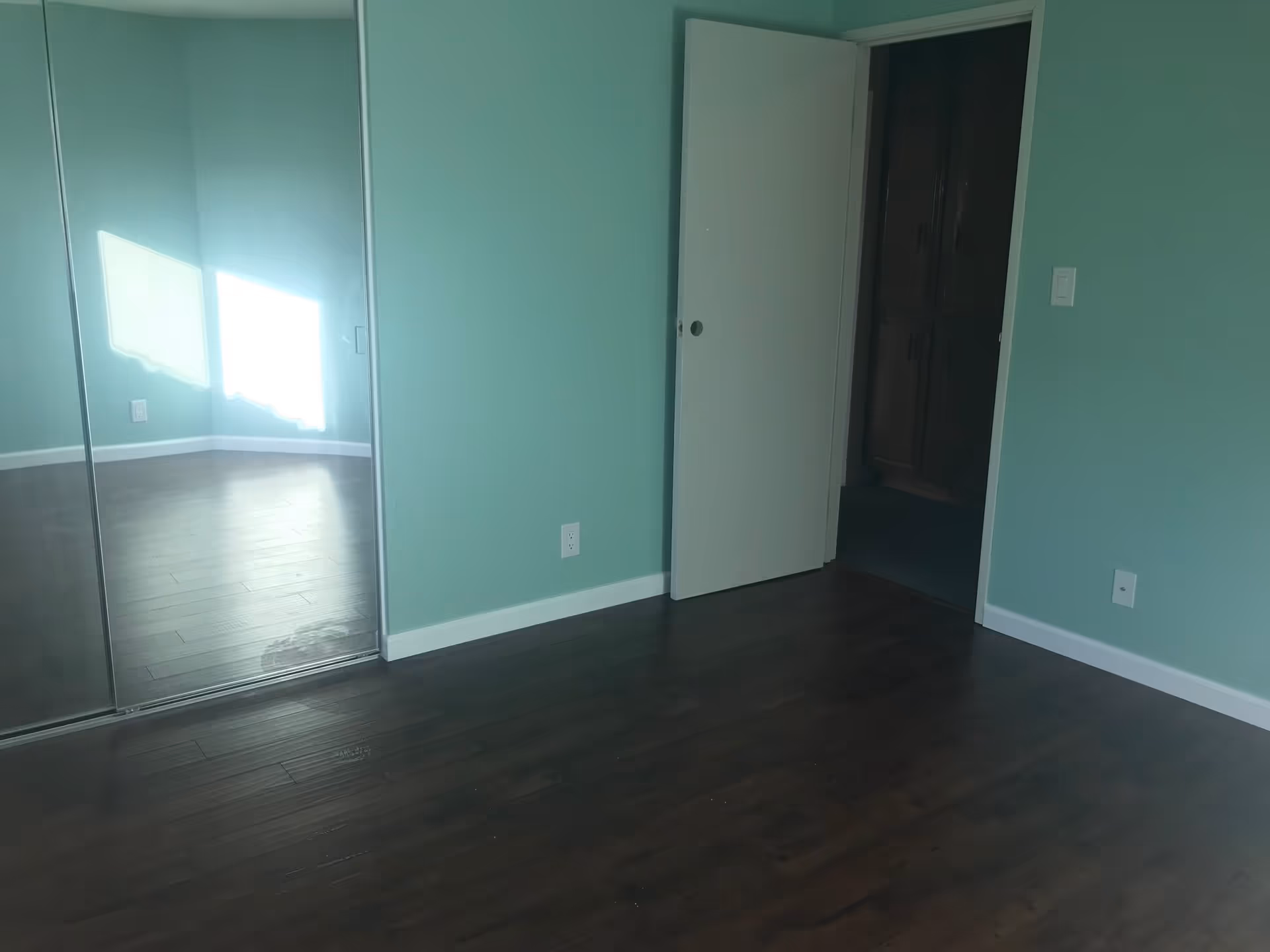 Empty room with teal walls, dark wooden floor, a sliding mirrored closet door reflecting sunlight from a window, and a partially open white door leading to another room.